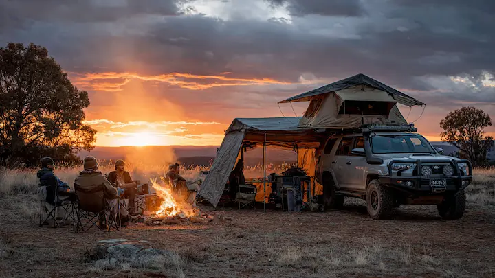 4x4 vehicles set up at a mountain campsite at sunset, one with a rooftop tent and awning, the other with a dome tent nearby and a campfire burning between them, overlooking a vast valley in the Australian high country.